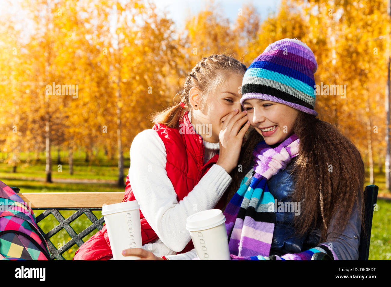 Two happy girls whispering 11 years old sitting on the bench in autumn park Stock Photo