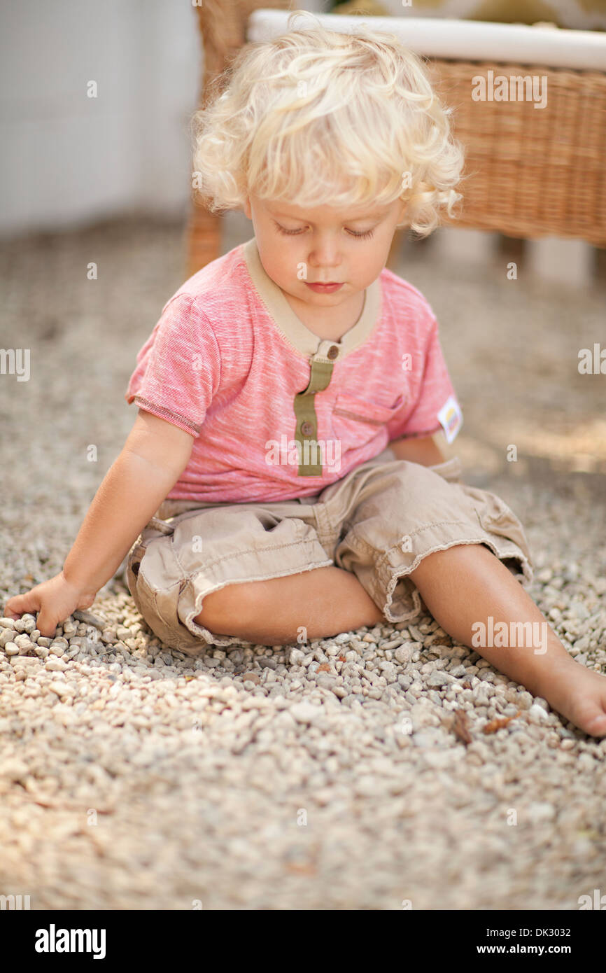 Blonde Toddler Boy With Curly Hair Playing In Pebbles On Patio