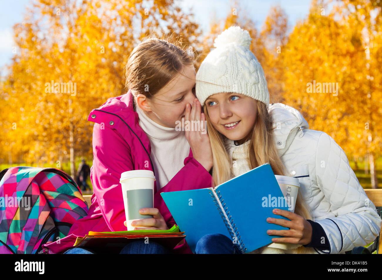 Two happy 14 years old girls whispering secrets and gossips in the autumn park sitting on the bench holding textbook and coffee Stock Photo