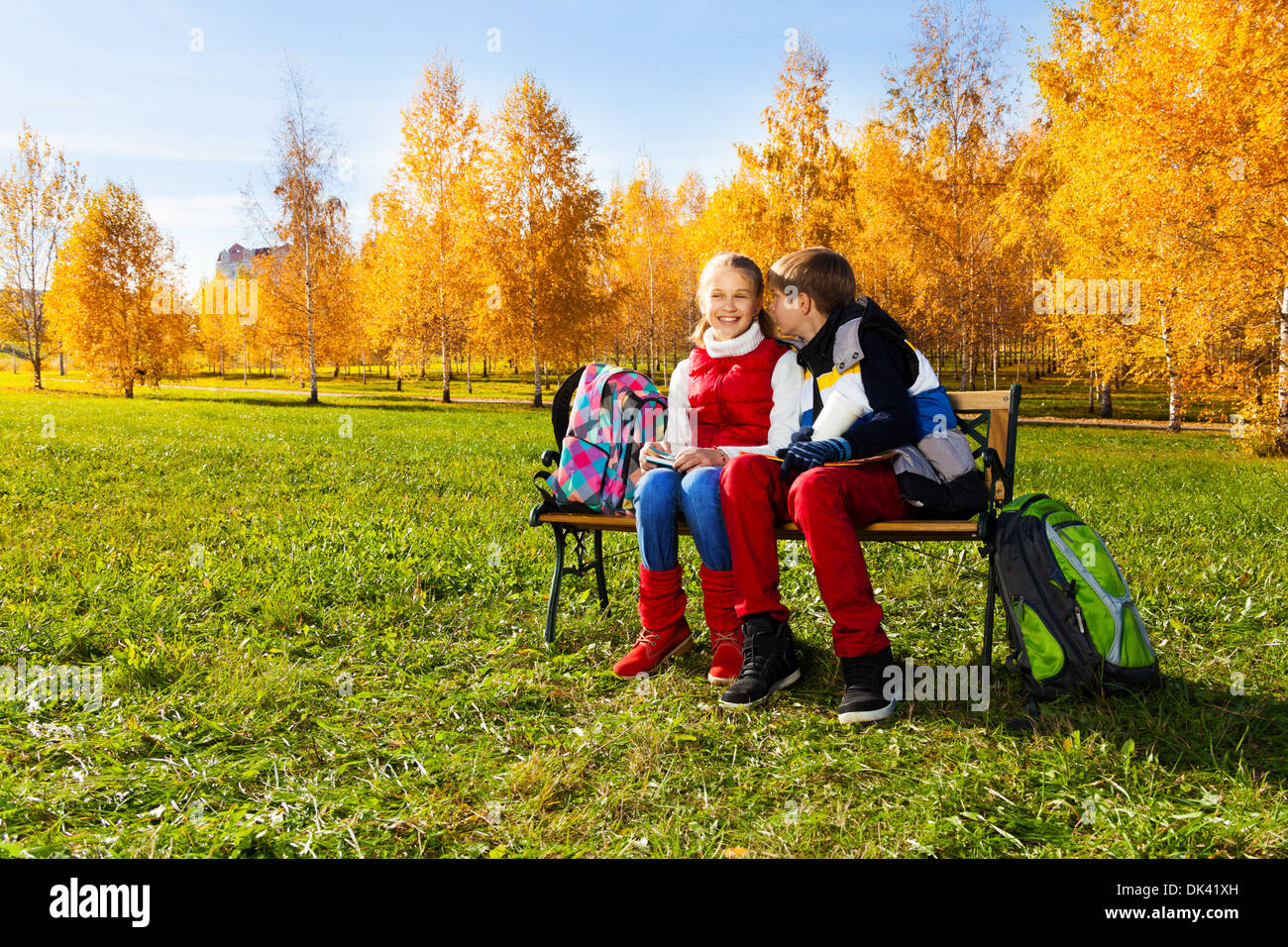 Couple children, boy whispering to girl sitting on the bench in autumn park with backpacks laying near by Stock Photo