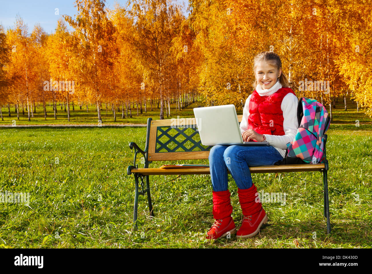 Happy blond 11 years old girl with amazing smile sitting on the bench with laptop doing homework outside in the autumn park on sunny day Stock Photo