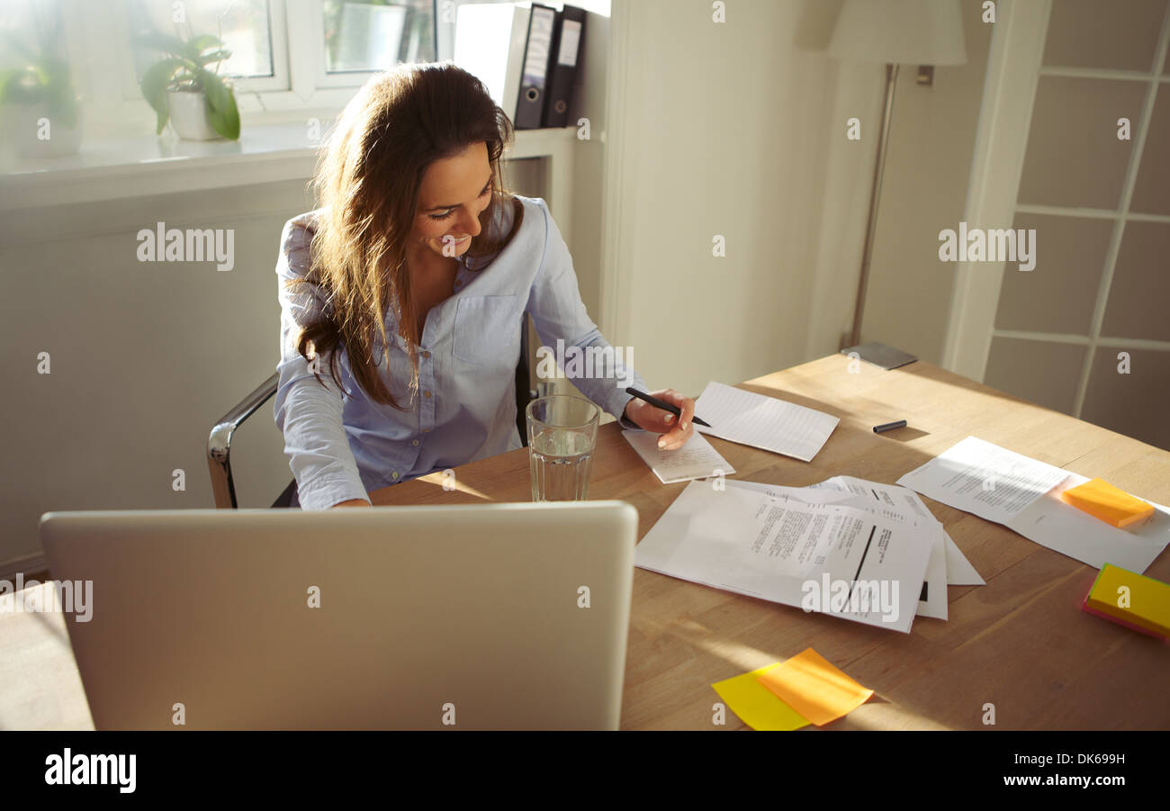 Pretty young female writing notes smiling. Beautiful caucasian female with laptop computer working from home office. Stock Photo