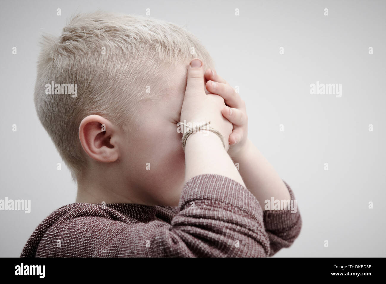 Portrait of boy wearing brown jumper, covering face with hands Stock Photo