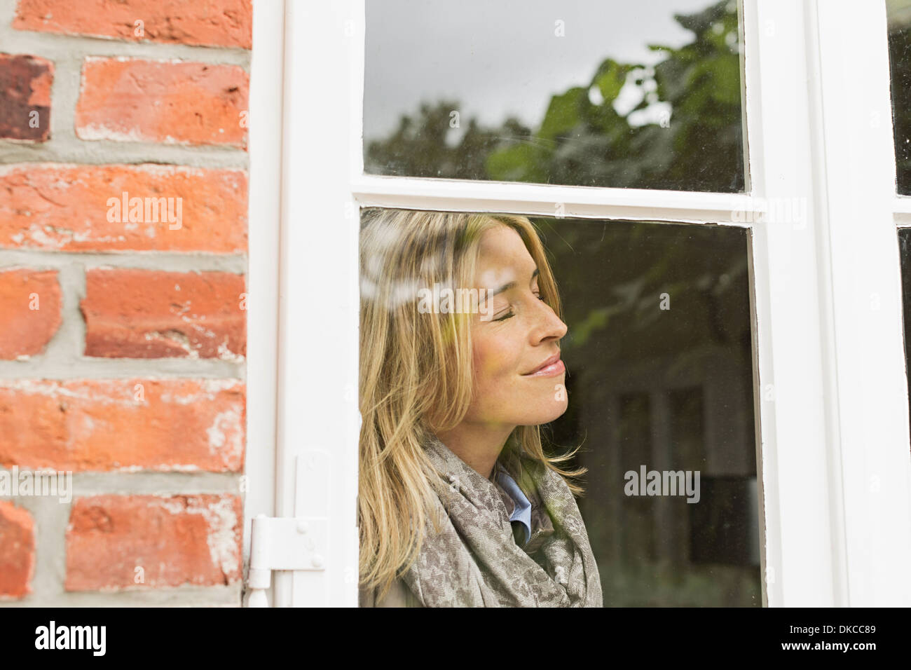 Mid adult woman by window with eyes closed, smiling Stock Photo