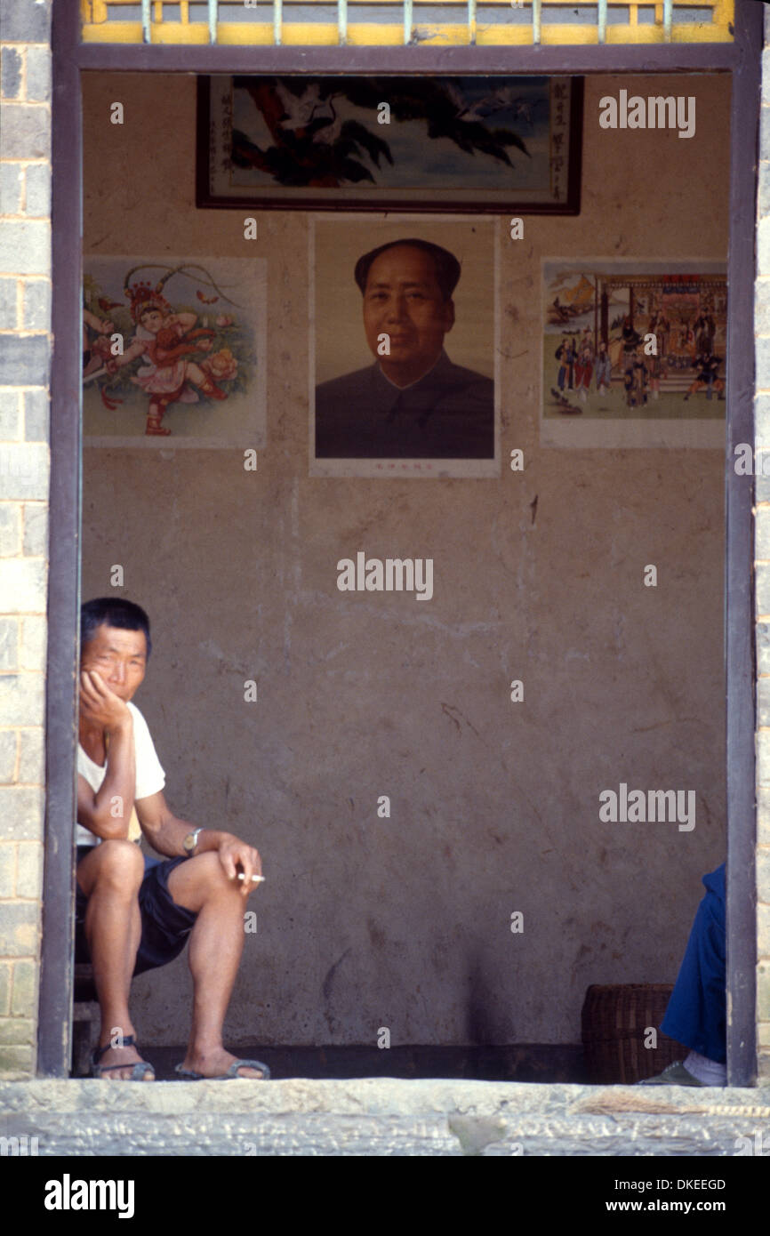 A man sitting at the entrance of his house, smoking. In the middle a poster of Mao Zedong was hang. Stock Photo