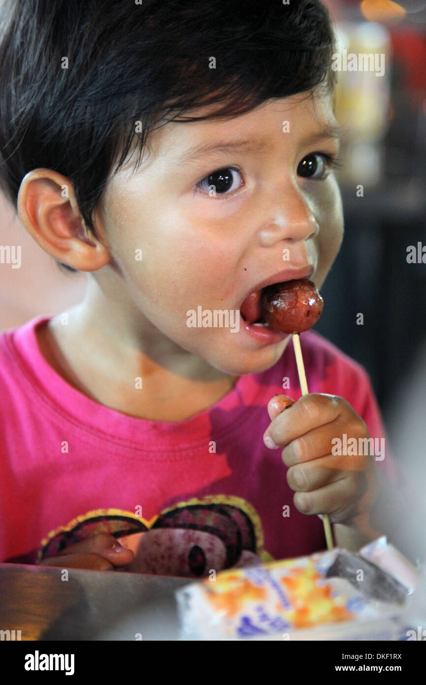 It's a photo of a Eurasian young boy who is eating a meat ball on a wood stick. He wears a Pink t-shirt Stock Photo