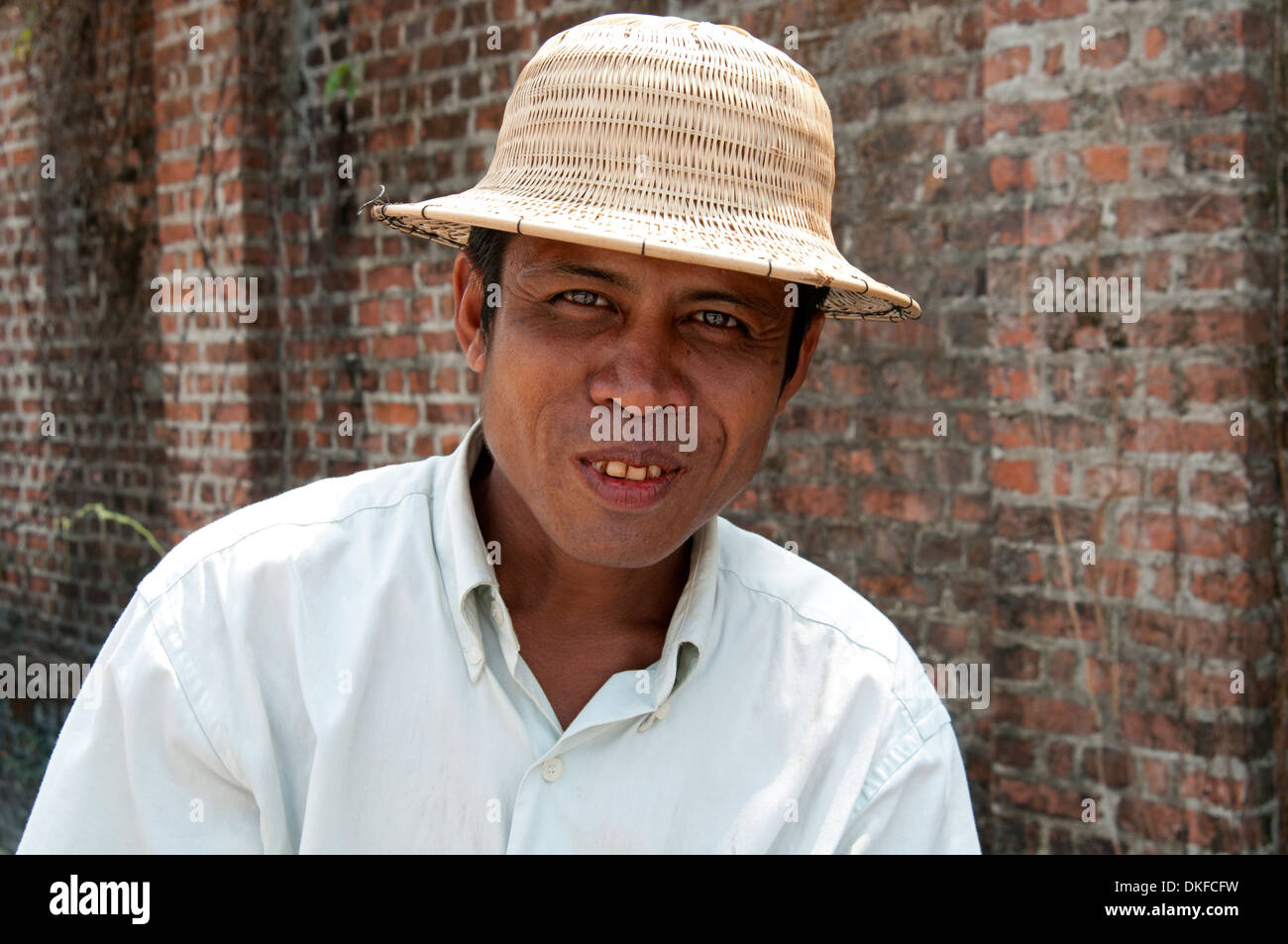Red stained betel nut grin on a Yangon bicycle taxi driver wearing a straw hat against a brick wall Myanmar (Burma) Stock Photo