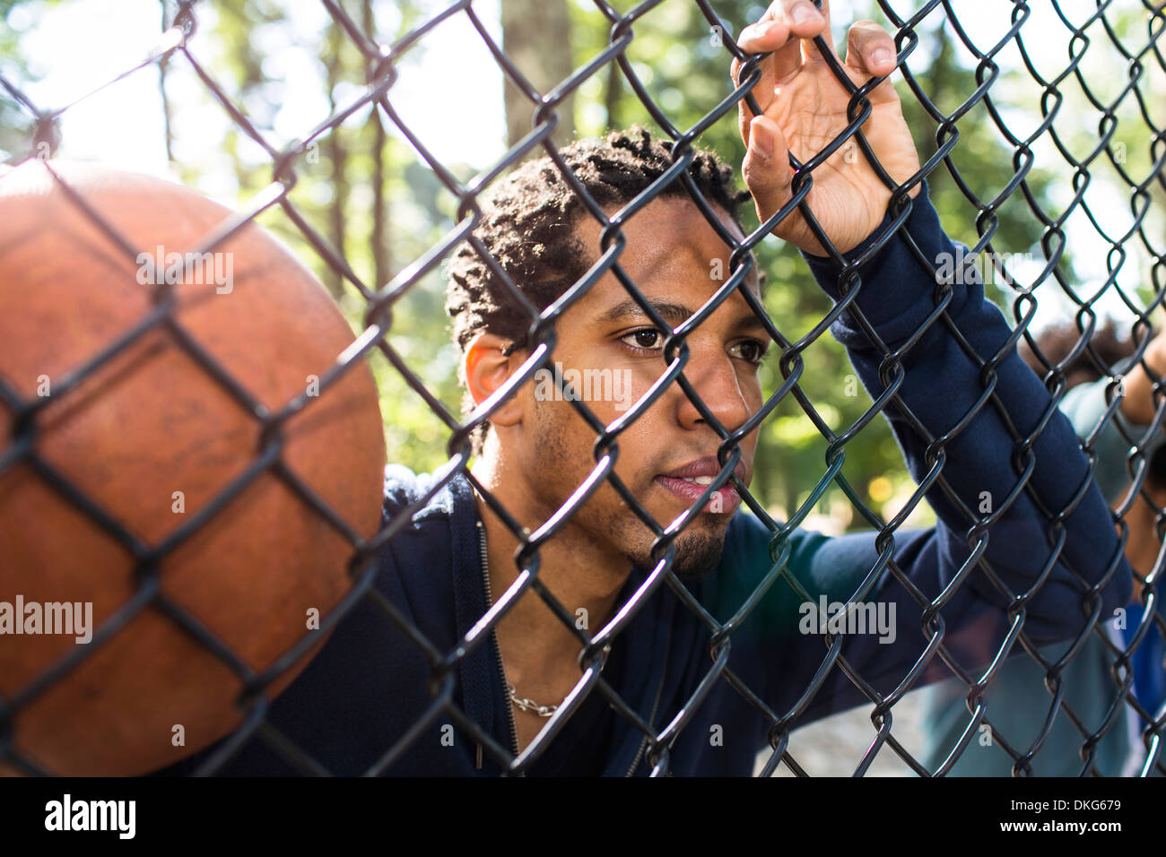 Portrait of young man holding basketball through wire fence Stock Photo