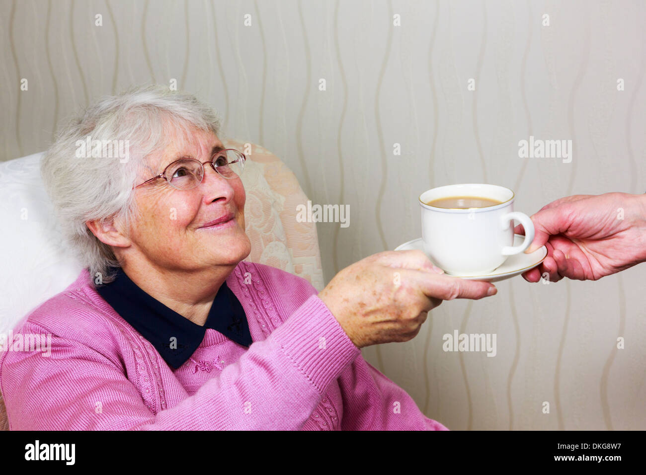Frail sweet happy old elderly senior woman looking and smiling at a caregiver handing her a cup of tea during daily help home visit. England UK Stock Photo