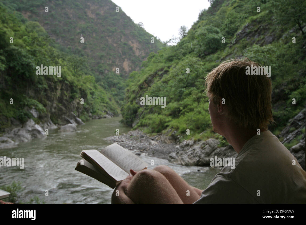 A young person reading a book by a slow flowing river, in the foothills of the Himalayas. Stock Photo