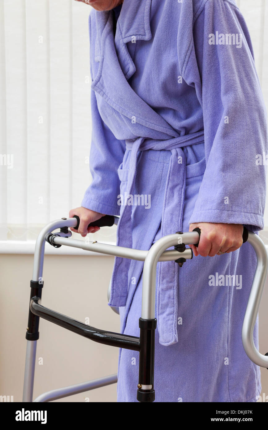 Elderly senior woman wearing a purple bathrobe holding using a zimmer frame walker or support trolley for walking in a home. England UK Britain Stock Photo