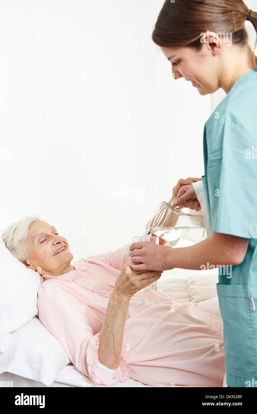 Geriatric nurse giving water to bedridden senior woman in nursing home