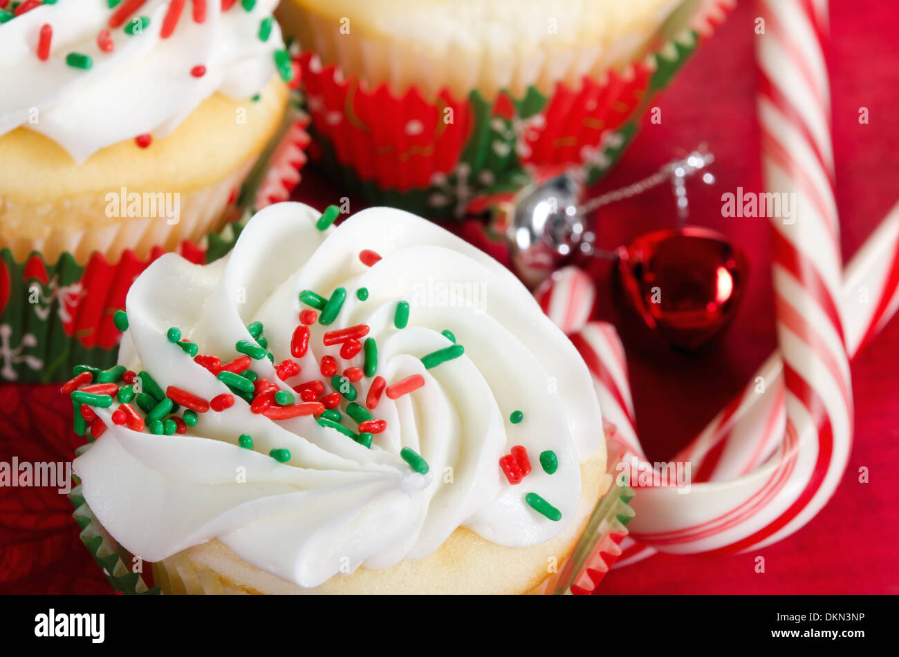 Holiday cupcakes with vanilla frosting and red and green sprinkles. Red holiday background with candy canes and bells. Stock Photo