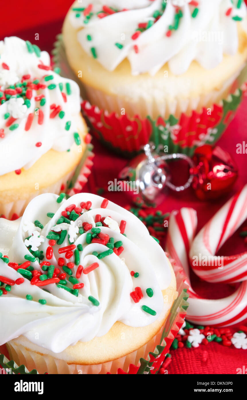 Holiday cupcakes with vanilla frosting and red and green sprinkles. Red holiday background with candy canes and bells. Stock Photo