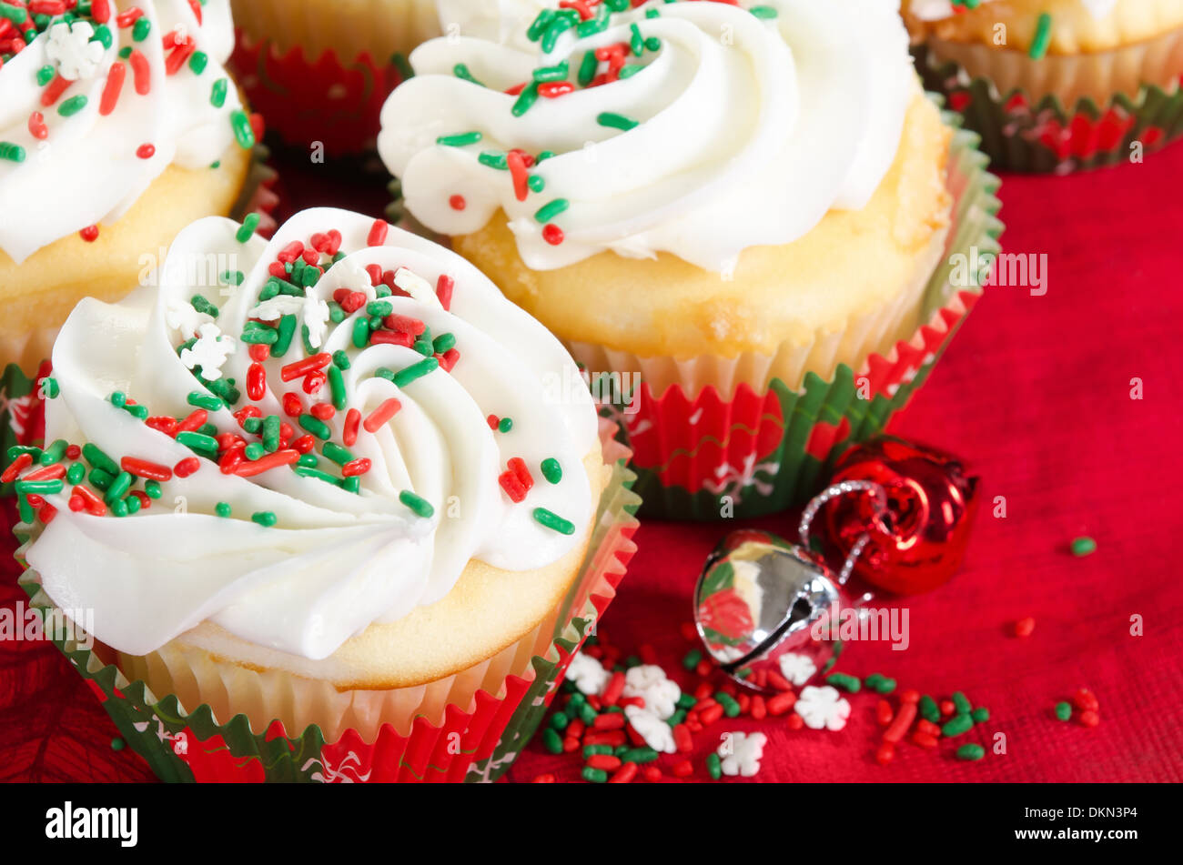 Holiday cupcakes with vanilla frosting and red and green sprinkles. Red holiday background with Christmas bells. Stock Photo