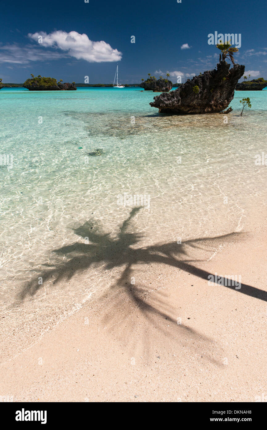 Shadow of a coconut palm tree on a tropical beach, a cruising yacht at anchor and views of limestone motus, small eroded islets in Fulaga lagoon. Fiji Stock Photo