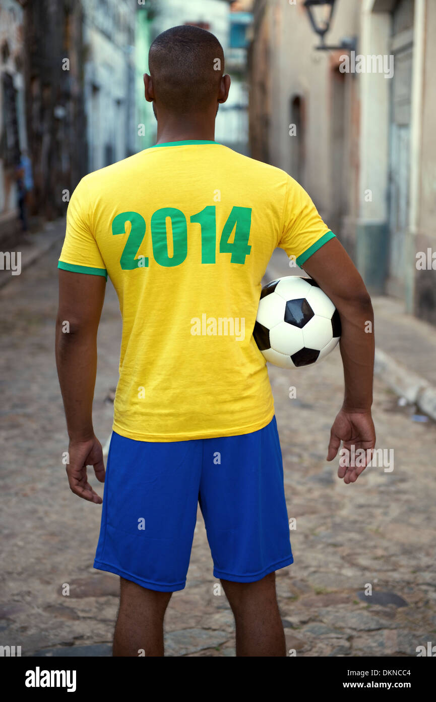 Brazilian football player in 2014 shirt stands holding a soccer ball on an old rustic village street Stock Photo