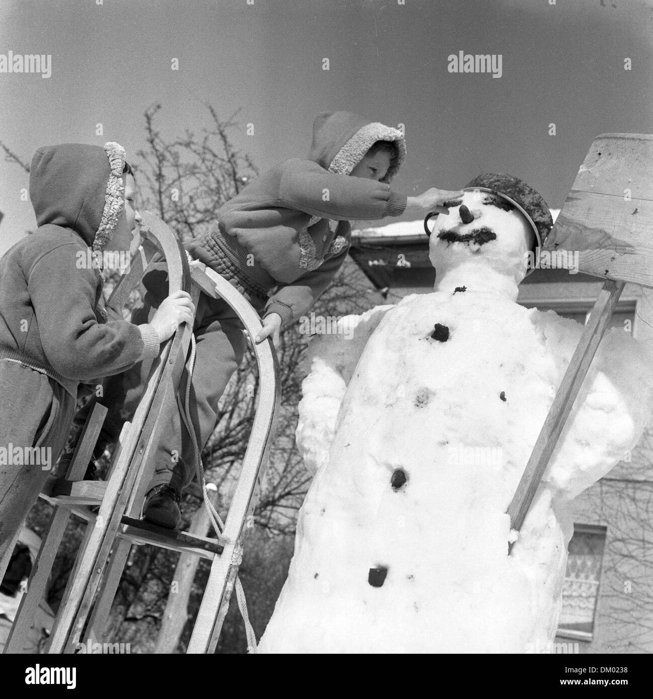 Children use a sledge as a ladder to place the carrot nose in the face of a snowman - playing in the snow, undated photograph from the early 1960ies. Photo: Heinz Junge Stock Photo