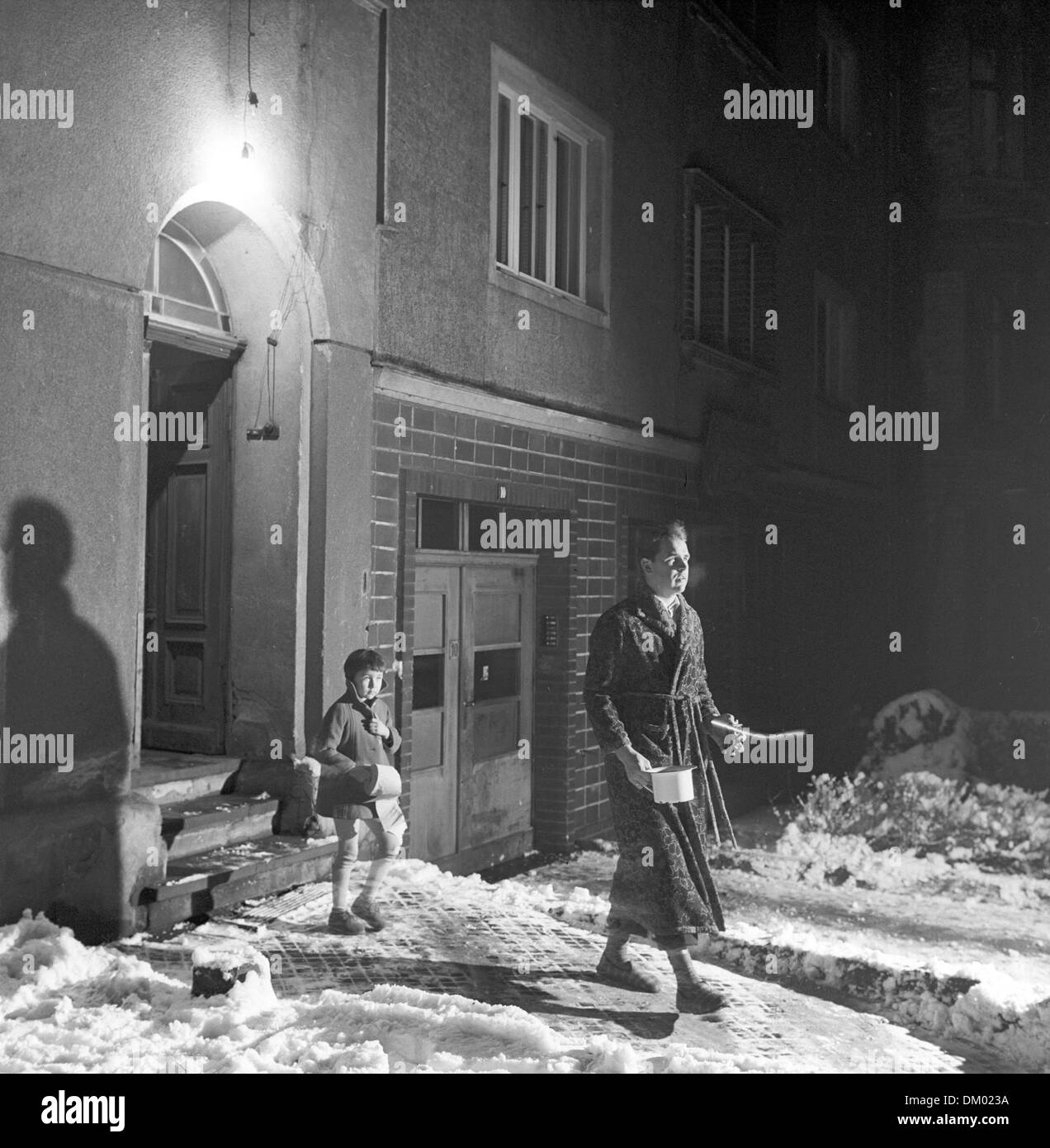 A small boy with a roll of toilet paper and a man with a dressing gown, flashlight, and a chamber pot step outside of an illuminated house . the toilet is frozen, undated photograph from the 1960ies. Photo: Fred Dahlberg Stock Photo