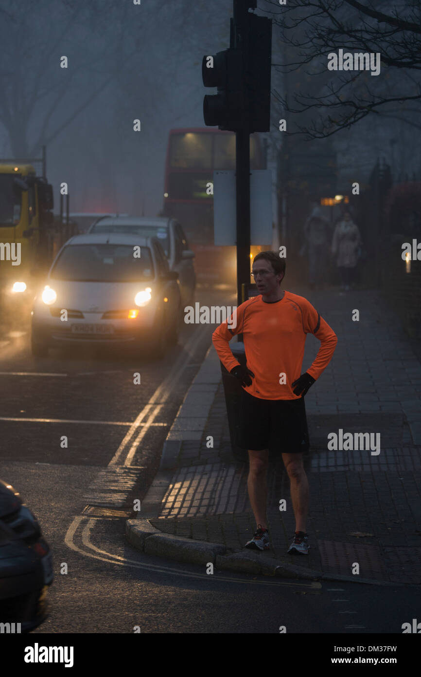 A jogger waits to cross in front of commuter traffic at dawn on a foggy morning in south London. Stock Photo