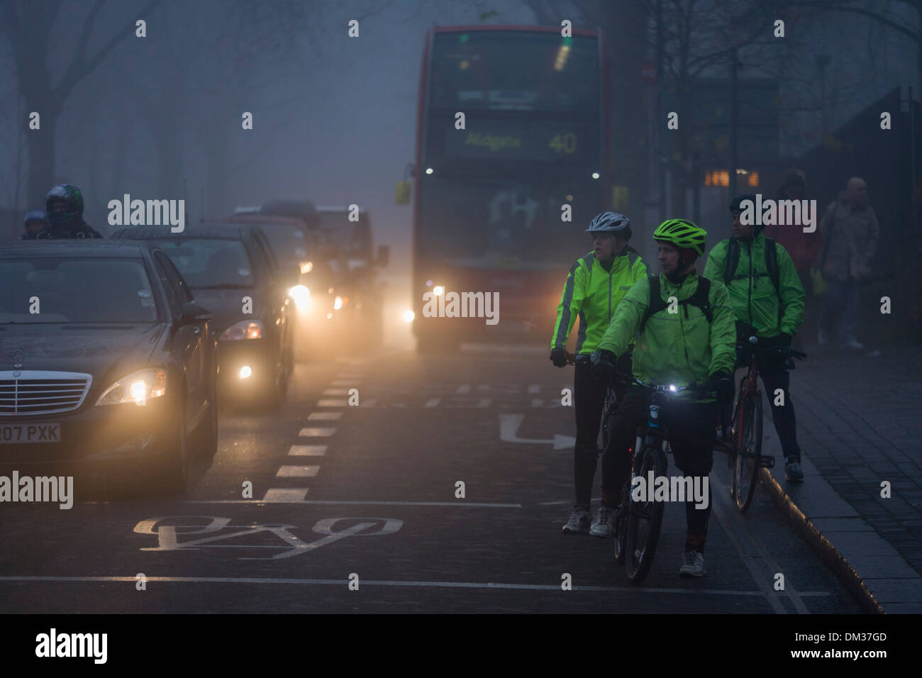 Commuting cyclists wait to cross a junction at dawn on a foggy morning in south London. Stock Photo