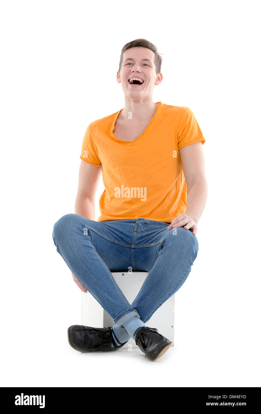 Funny young man, wearing a yellow T-shirt and slim jeans, laughing out loud while sitting on a cube, isolated Stock Photo