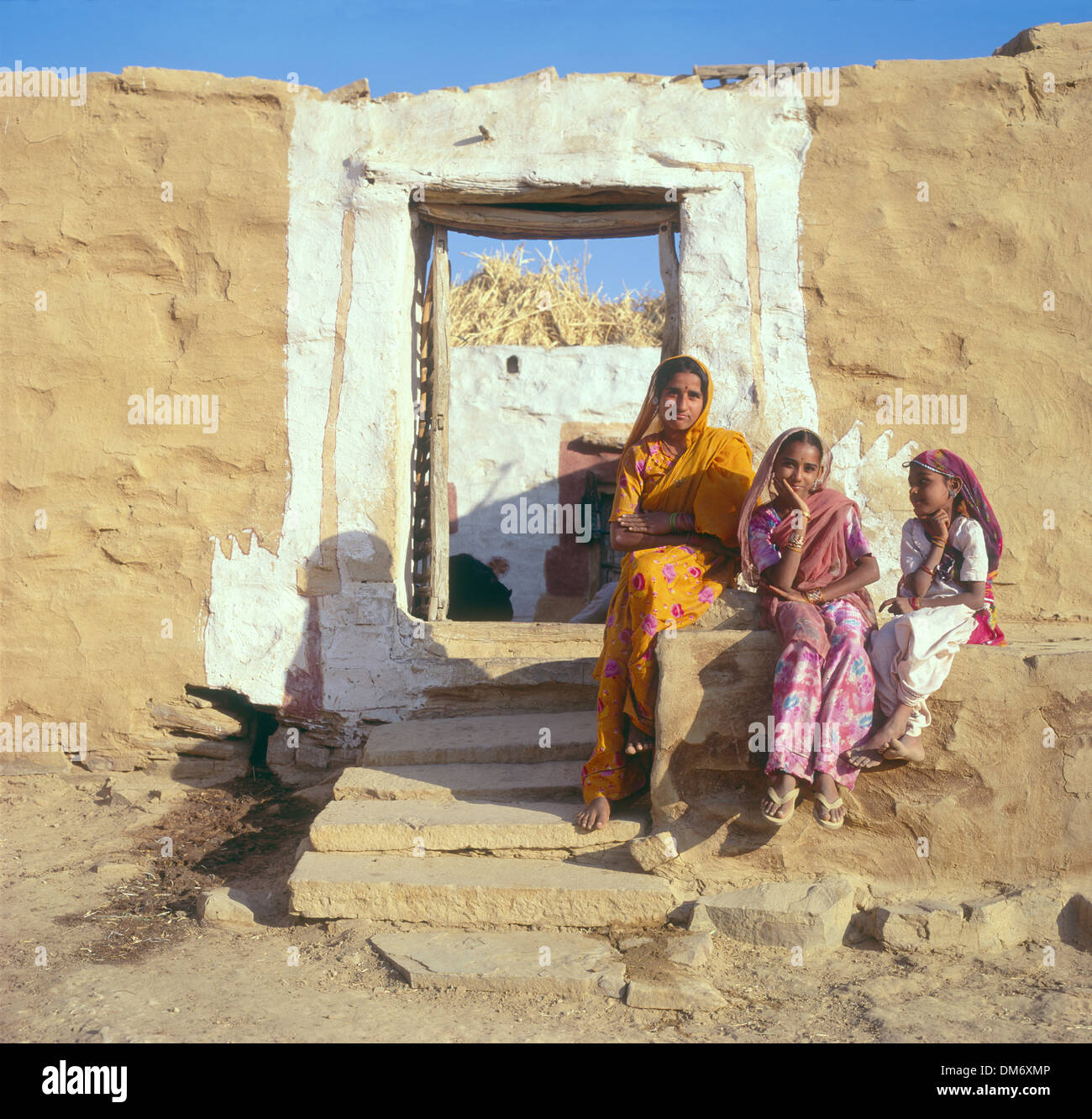 Woman and two girls in Saris outside an adobe house in desert Village outside Jaisalmer, Rajasthan, India, sand coloured walls Stock Photo