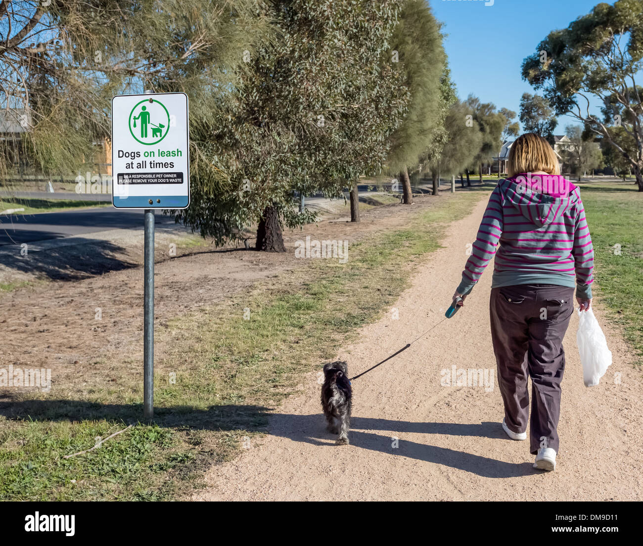 Senior lady carrying dog dropping bag walking puppy in park past responsible dog ownership sign. Stock Photo