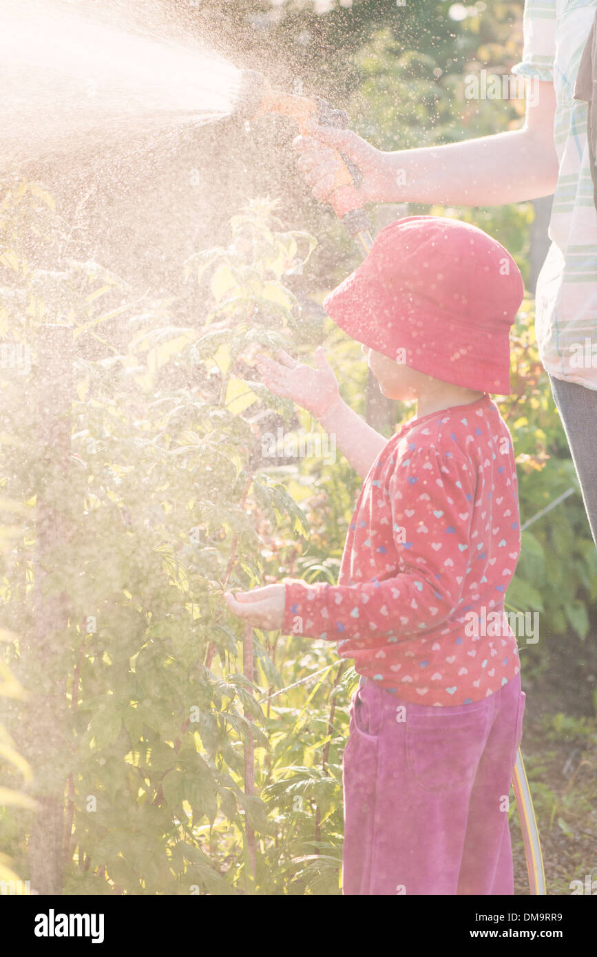 Lifestyle summer scene. Little girl playing with parent in garden, feeling water from sprinkler Stock Photo