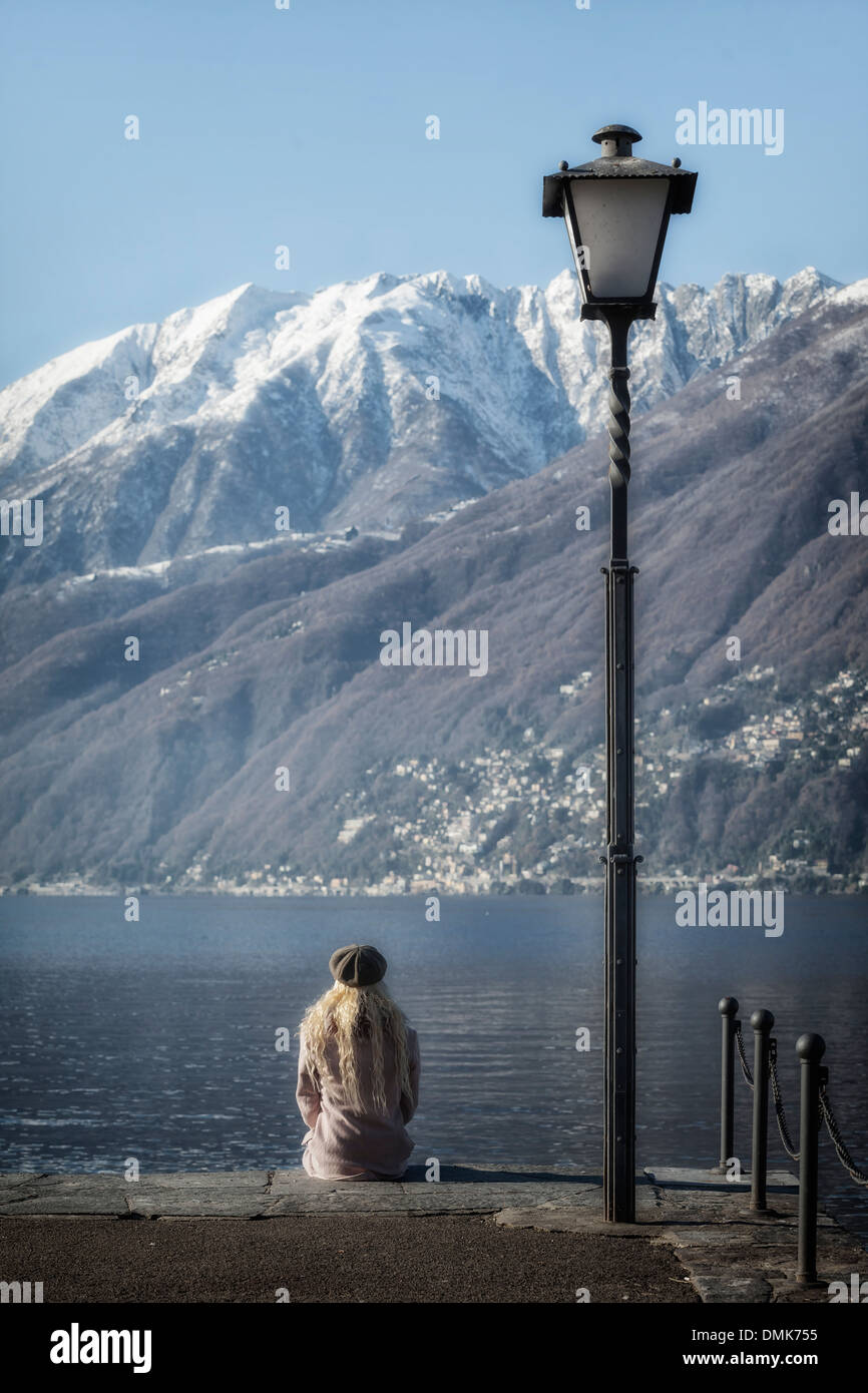 a girl in a pink coat is sitting at a lake, in the background snow covered mountains Stock Photo