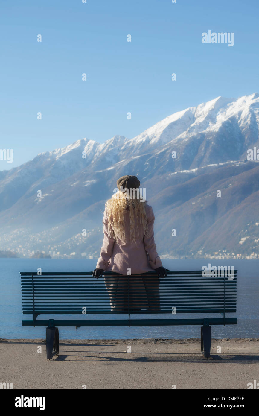 a girl in a pink coat is sitting on a bench at a lake Stock Photo