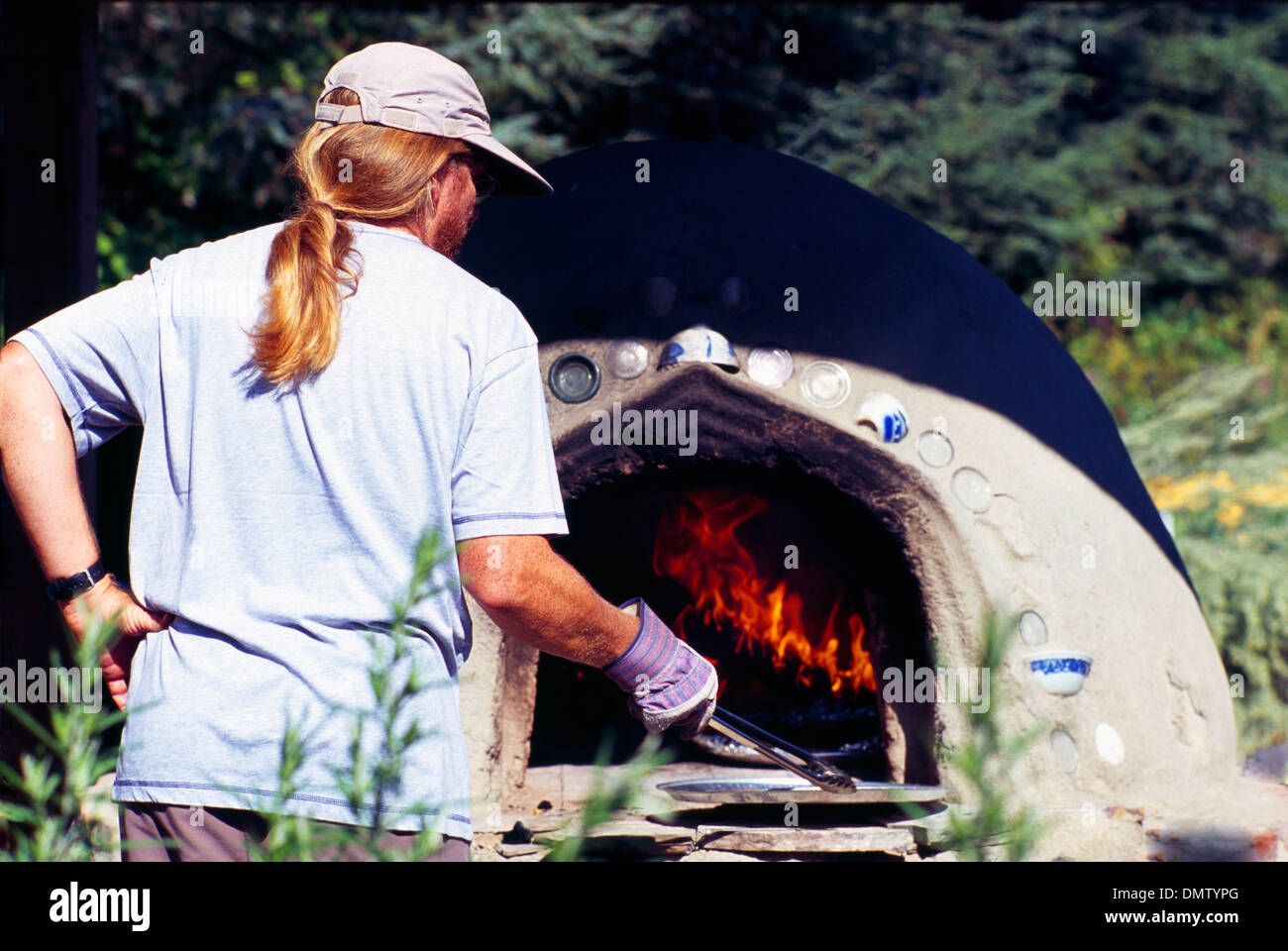 Working Man with a Long Ponytail baking Garlic Bread in Outdoor Wood Fired Cob Oven at Garlic Festival Stock Photo