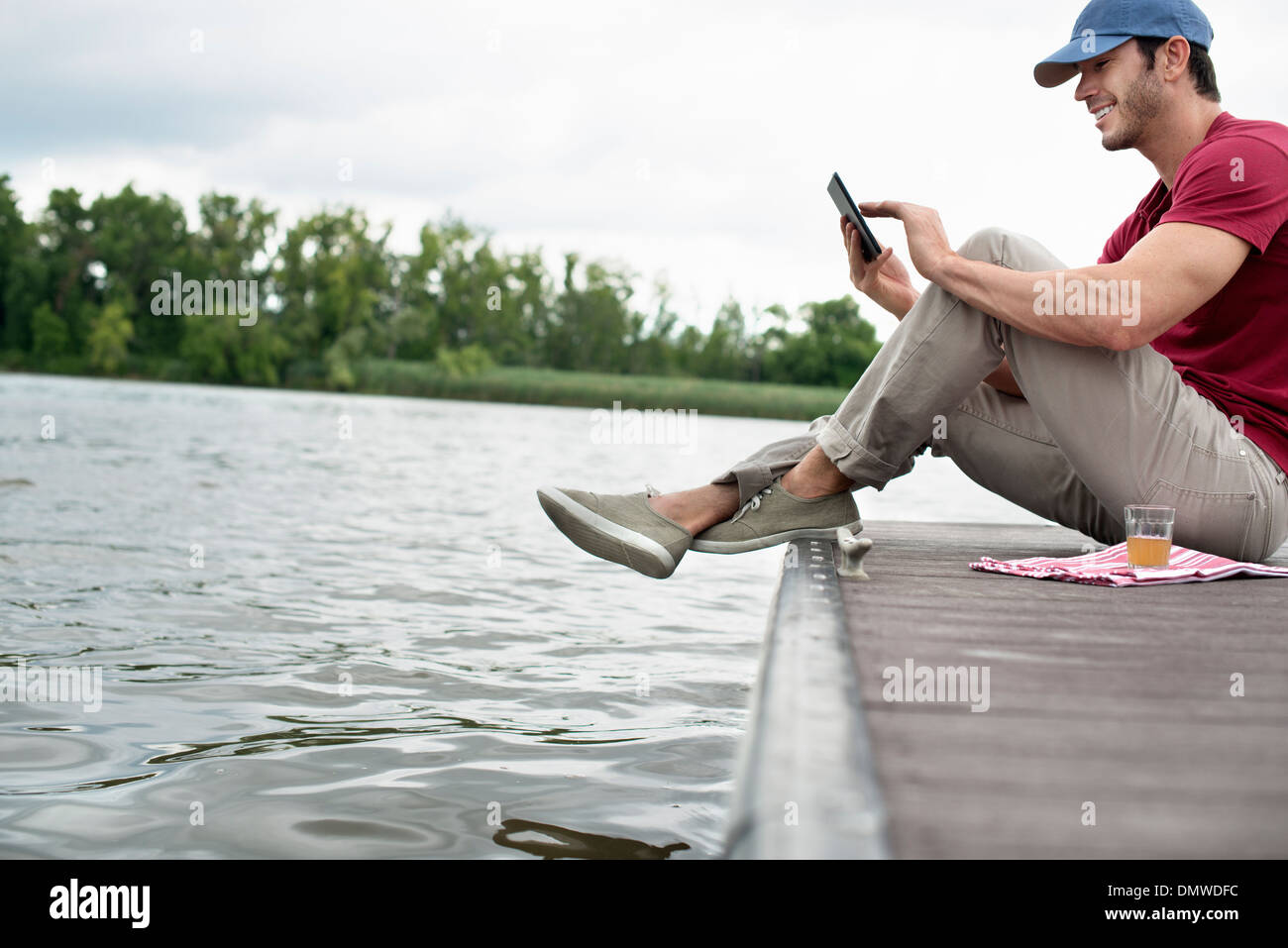 A man seated on a jetty by a lake using a digital tablet. Stock Photo