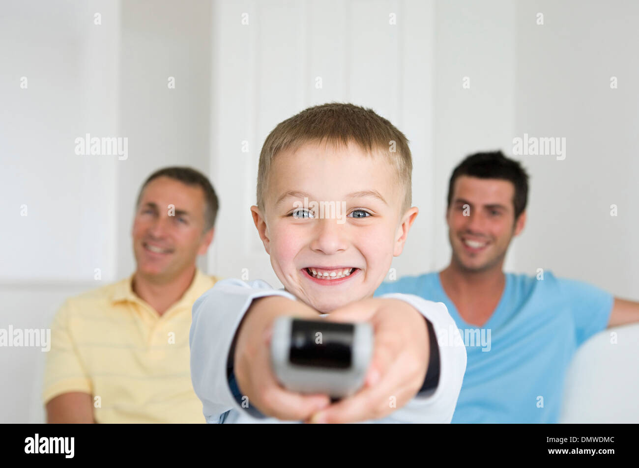 A child a boy holding a television remote control. Two men on  sofa behind him. Stock Photo