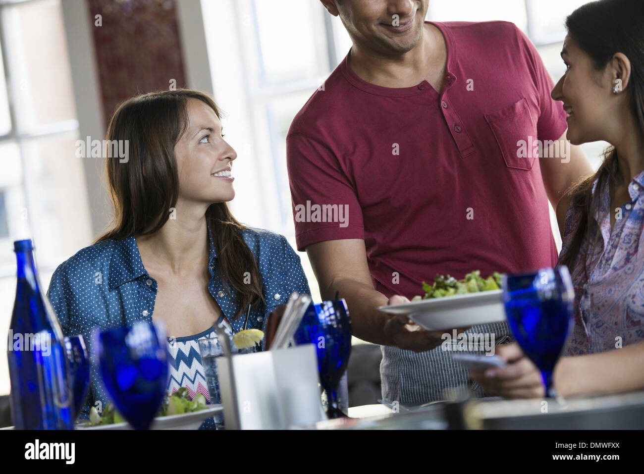 A cafe interior. A man in a waiter's apron serving a meal to two women at a table. Stock Photo