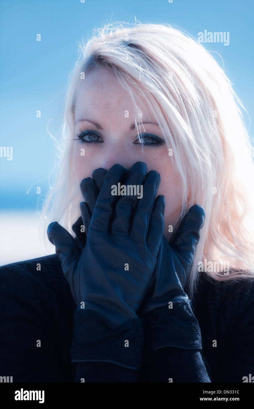 portrait of a beautiful young woman with long blond hair who is holding her hands with black gloves in front of her mouth Stock Photo
