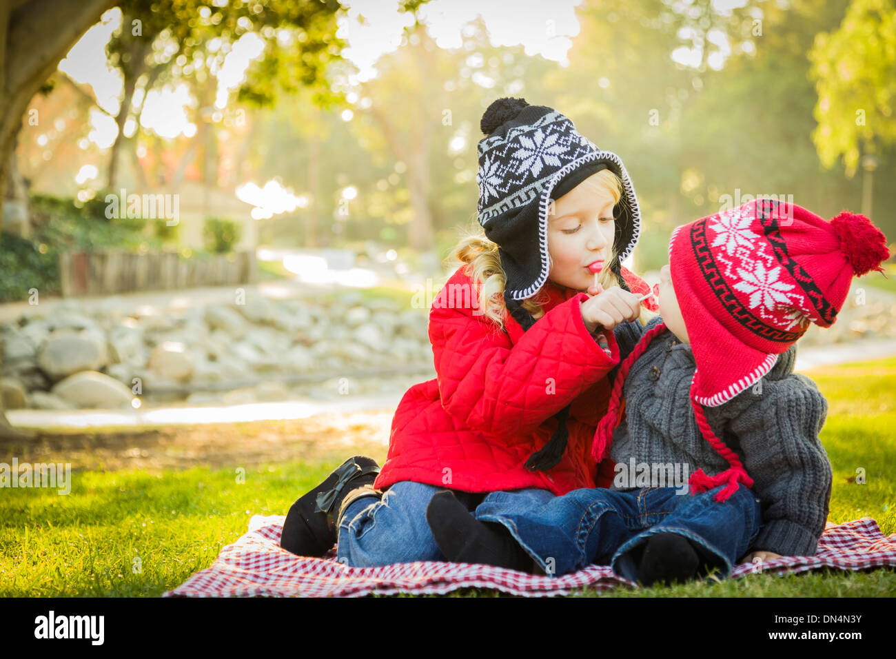 Little Girl with Her Baby Brother Wearing Winter Coats and Hats Sharing a Lollipop Outdoors at the Park. Stock Photo