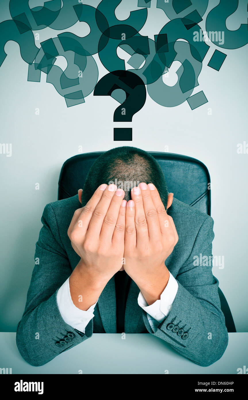 a businessman sitting in a desk with his hands in his head and a cloud of question marks above him Stock Photo
