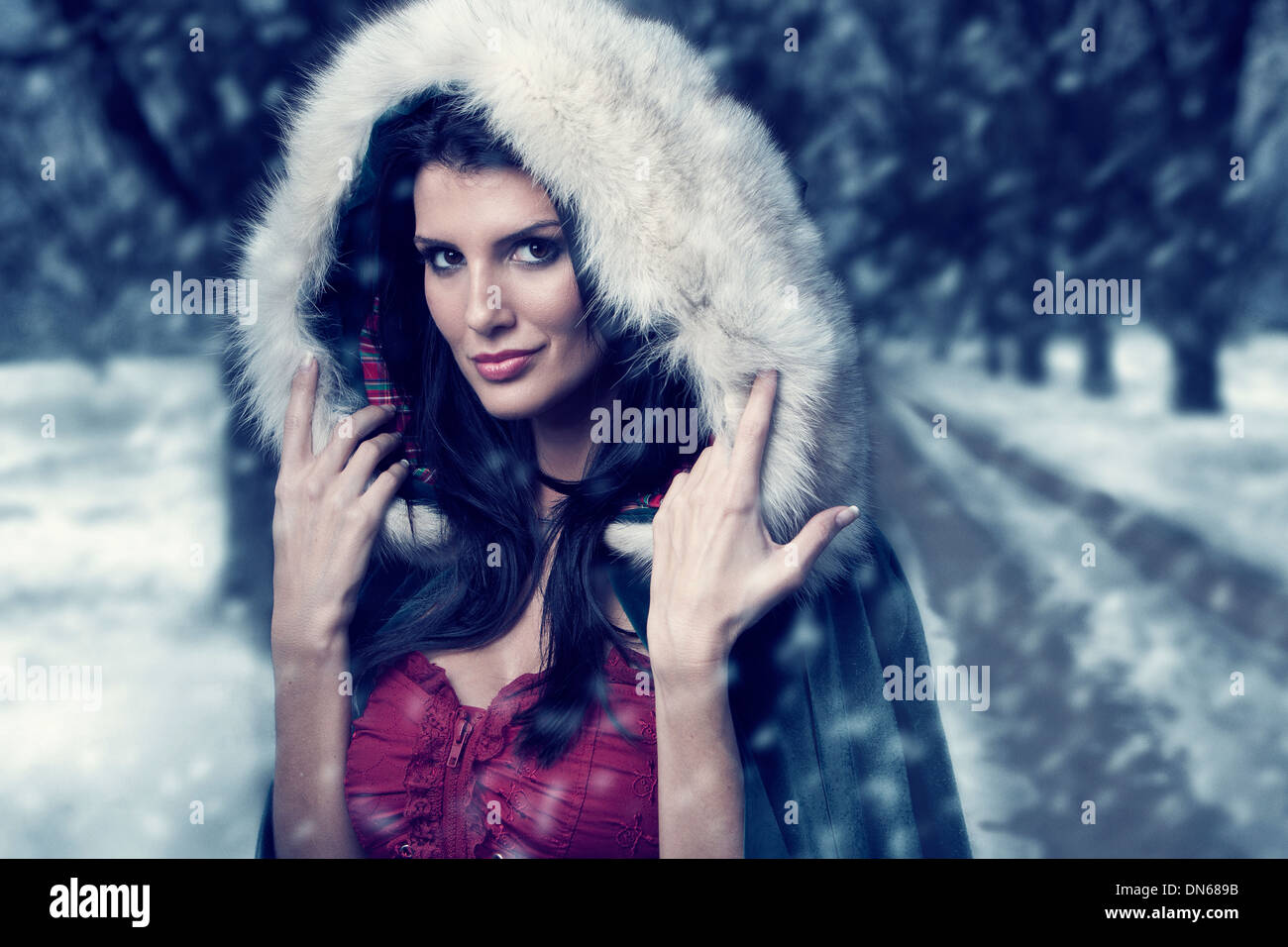 Woman in cape with hood standing on wooded path in the snow Stock Photo