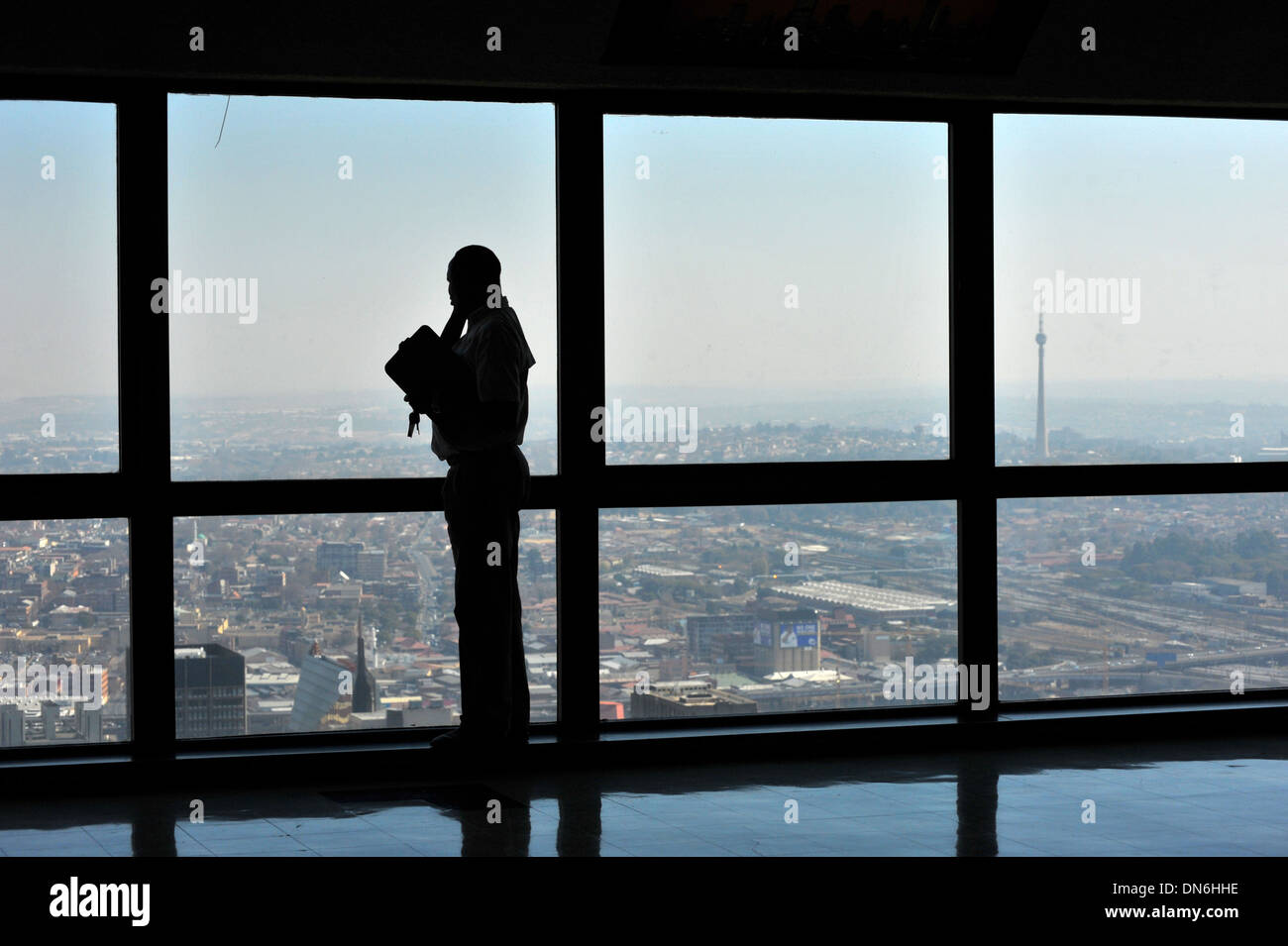 A male silhouetted against full length windows looking out onto a city from the top of a skyscraper. Stock Photo