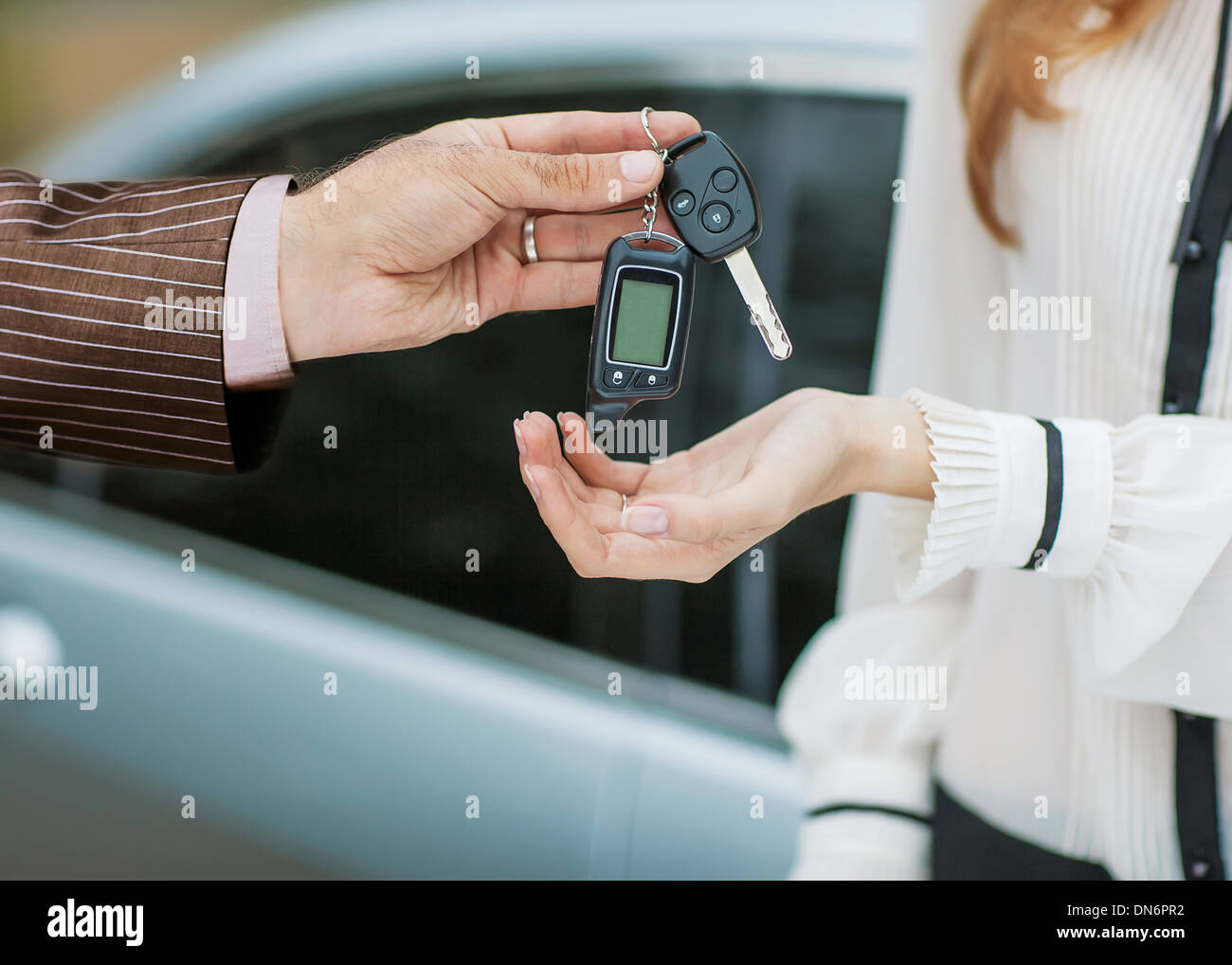 Male hand giving car key to female hand. She is holding a cell phone. In the background, a fragment of the car. Stock Photo