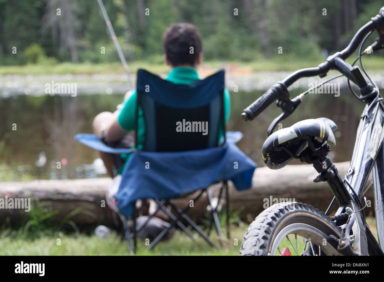 A teenage boy fishing by the lakeside in his camp chair. Focus on the bike. Stock Photo