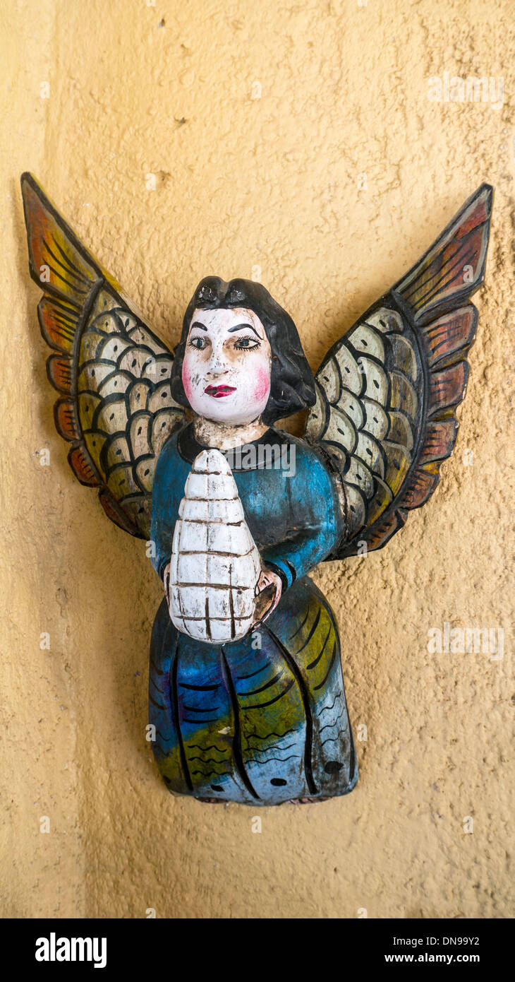 brightly colored charming crudely painted Mexican folk art bas relief of angel with golden wings hanging on yellow wall Mexico Stock Photo