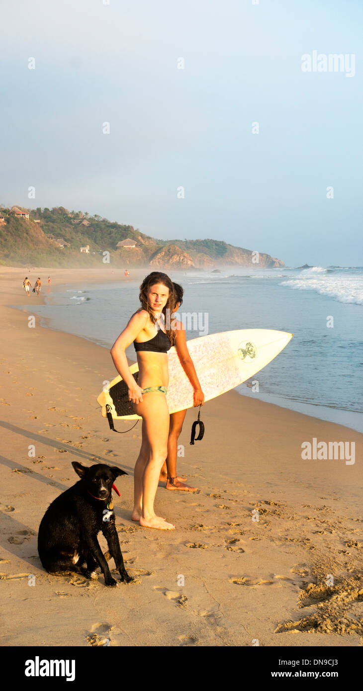 2 lithe surfer girls in bikinis with surf boards & adorable black mutt as sun is setting over beach & sea San Agustinillo Mexico Stock Photo