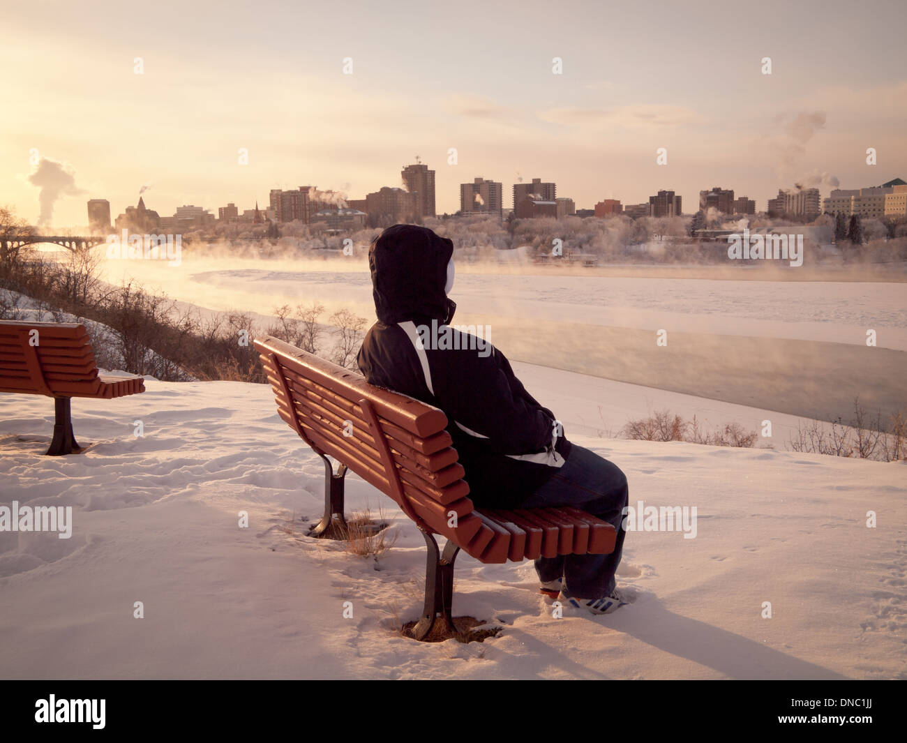 A man sits on a park bench overlooking the South Saskatchewan River on a cold, winter day in Saskatoon, Saskatchewan, Canada. Stock Photo