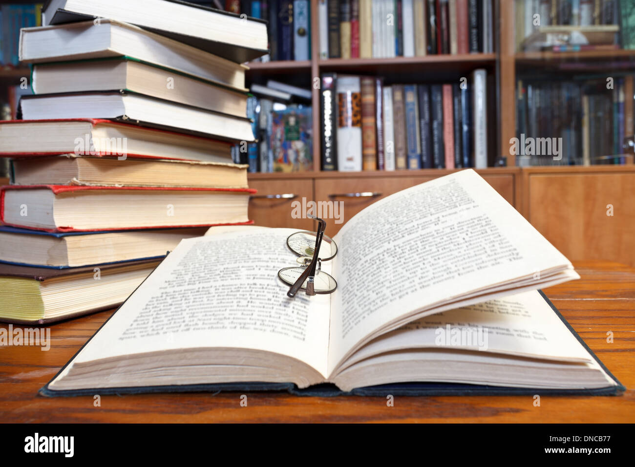 stack of books and open book and round glasses close up on wooden table near bookcases Stock Photo