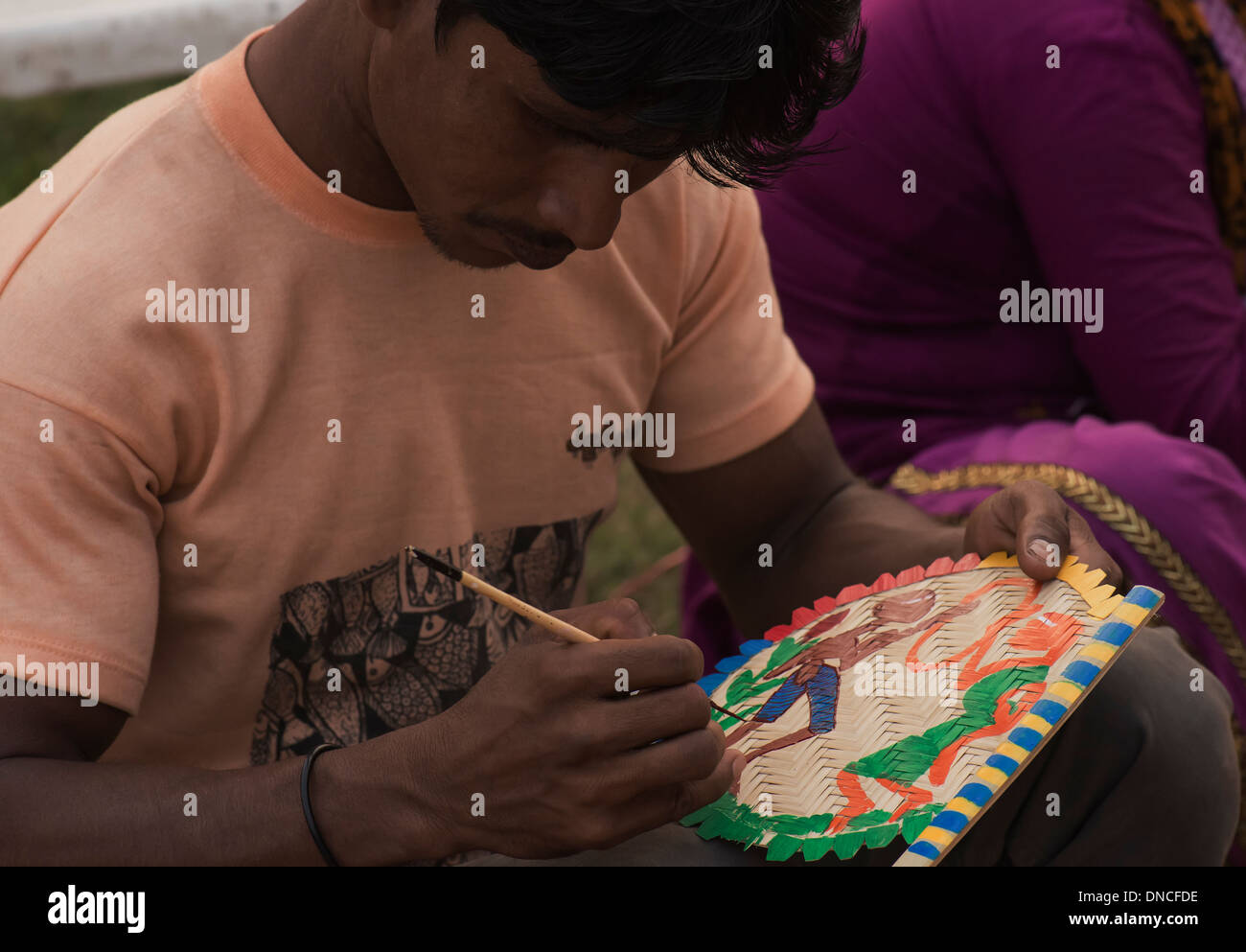 Indian male craft person paints on Bamboo hand fan the sketch in color of a boy and girl dancing. Stock Photo