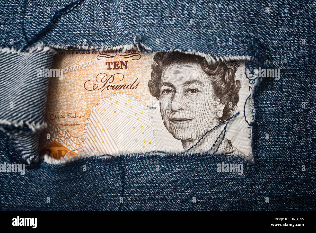 Ten pounds bill through torn blue jeans texture. British economy concept. Stock Photo