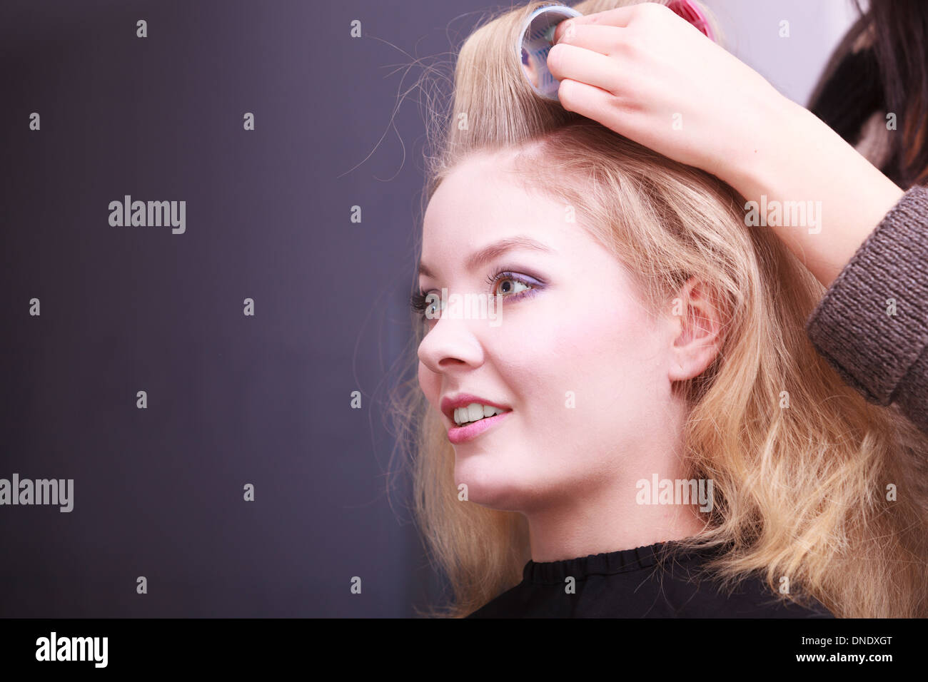 Beautiful young woman in beauty salon. Blond girl with hair curlers rollers by hairdresser. Hairstyle. Stock Photo