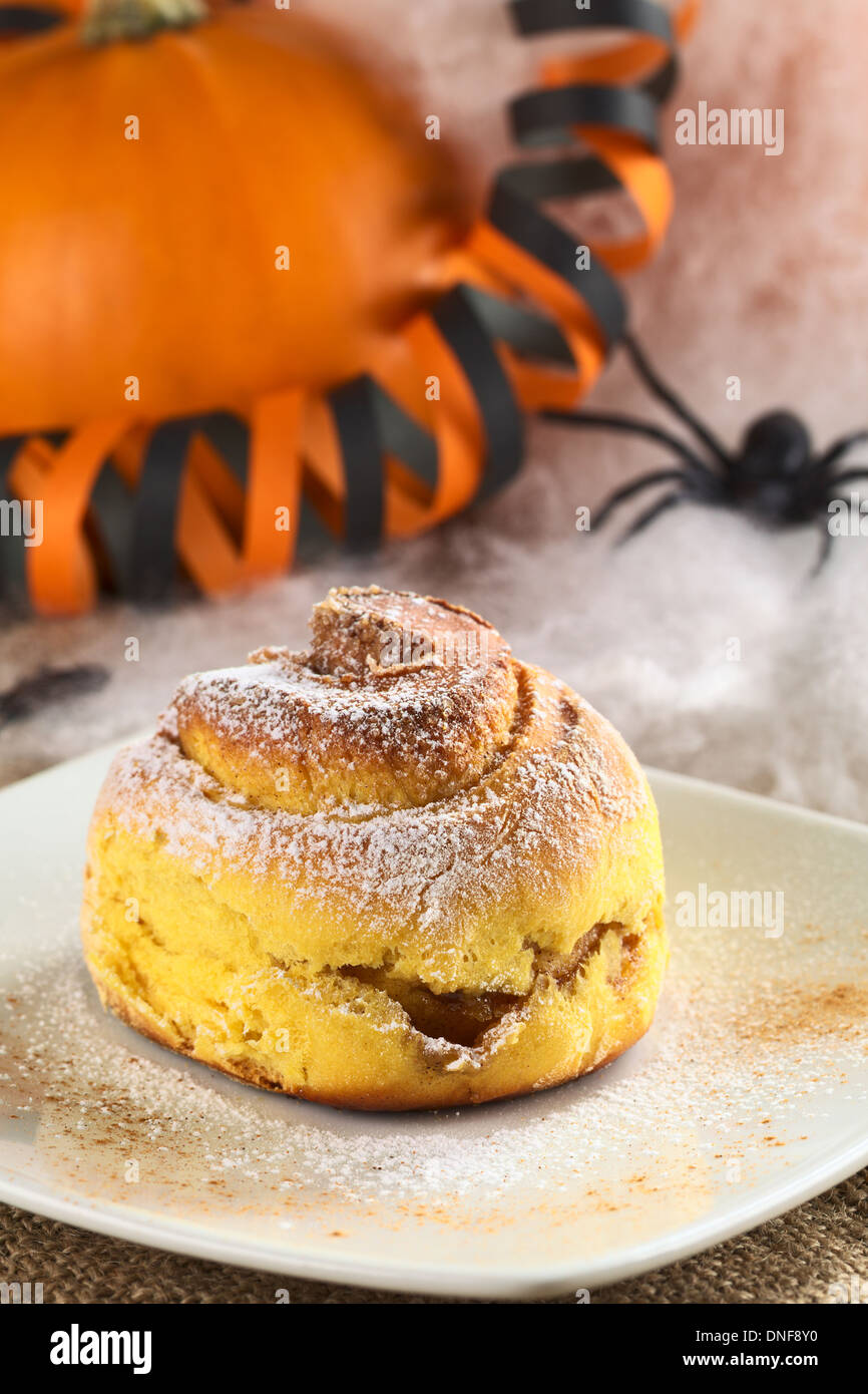 Pumpkin cinnamon roll garnished with confectioner's sugar and cinnamon, Halloween decoration in the back Stock Photo